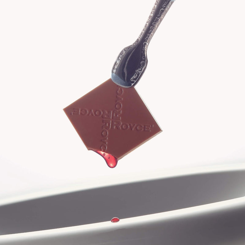 ROYCE' Chocolate - Prafeuille Chocolat Berry Cube - Image shows a silver tong holding a brown chocolate square with red sauce and engraved with the words ROYCE. Accents include a white plate-like surface. Background is in color white.