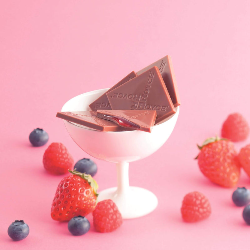 ROYCE' Chocolate - Prafeuille Chocolat Berry Cube - Image shows brown chocolate squares with red sauce and engraved with the words ROYCE, as placed in a white glass. Accents include red strawberries and raspberries and dark blue blueberries. Background is in pink color.