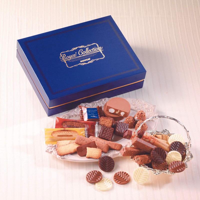 ROYCE' Chocolate - ROYCE' Collection Blue - Image shows on top left a blue box. Gold text says ROYCE' Collection ROYCE'. Below the box are a mix of confections in various shapes and colors, as arranged in accents such as a glass plate and silver bowl. Background and surface is a white tablecloth.
