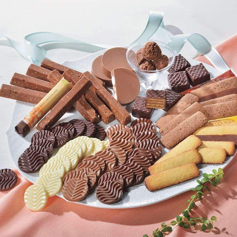 ROYCE' Chocolate - ROYCE' Collection Blue - Image shows a white plate and clear glass filled with various confections and chocolates in different shapes and colors. Accents include a white ribbon on top, a peach-colored table cloth, a white-colored surface on top, and some green leaves.
