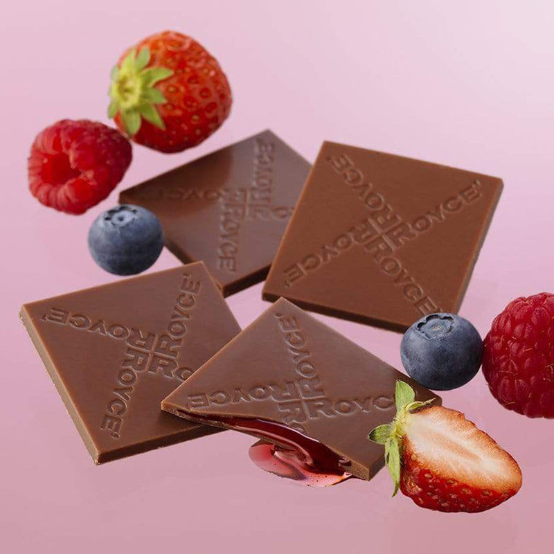 ROYCE' Chocolate - Prafeuille Chocolat Berry Cube - Image shows brown chocolate squares with red sauce and engraved with the words ROYCE. Accents include red strawberries and raspberries and dark blue blueberries. Background is in pink color.