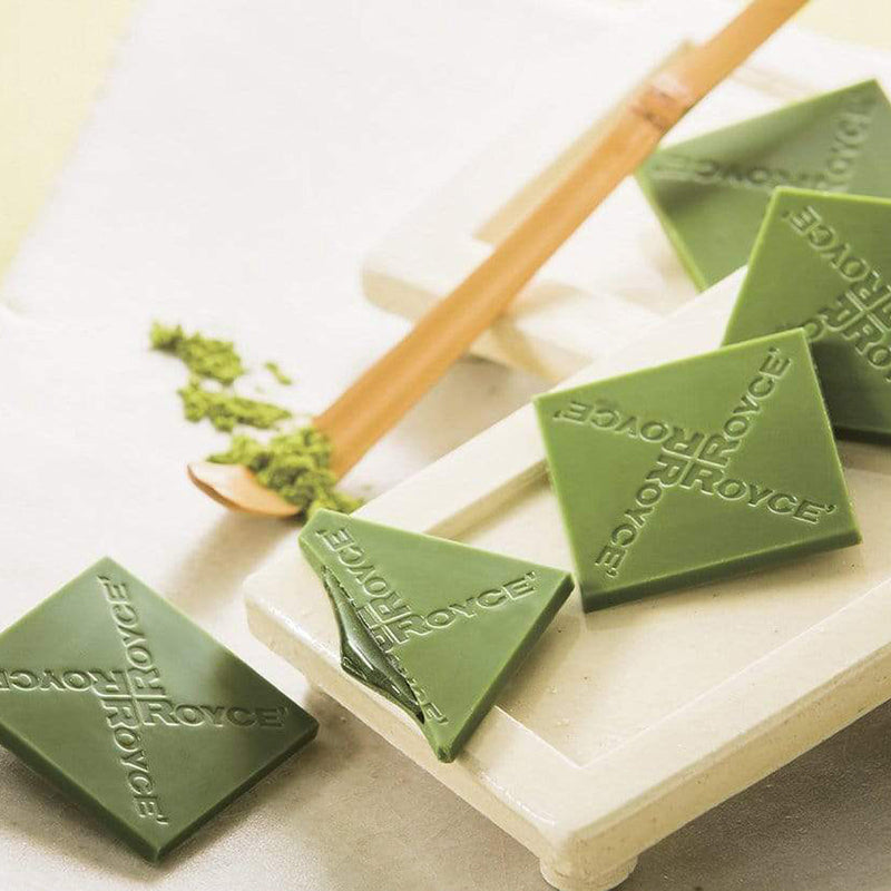 ROYCE' Chocolate - Prafeuille Chocolat Matcha - Image shows green chocolate squares filled with green sauce and engraved with the words ROYCE, as placed on white trays. Accents include a yellow wooden stick with green tea powder. Background is in white color.