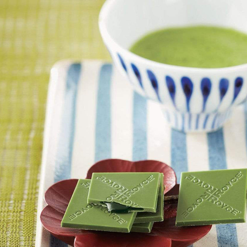 ROYCE' Chocolate - Prafeuille Chocolat Matcha - Image shows green chocolate squares filled with green sauce and engraved with the words ROYCE, as placed on a brown flower-shaped plate and a plate with blue and white stripes. Accents include a tea cup in blue and white filled with green tea. Background is in green color.