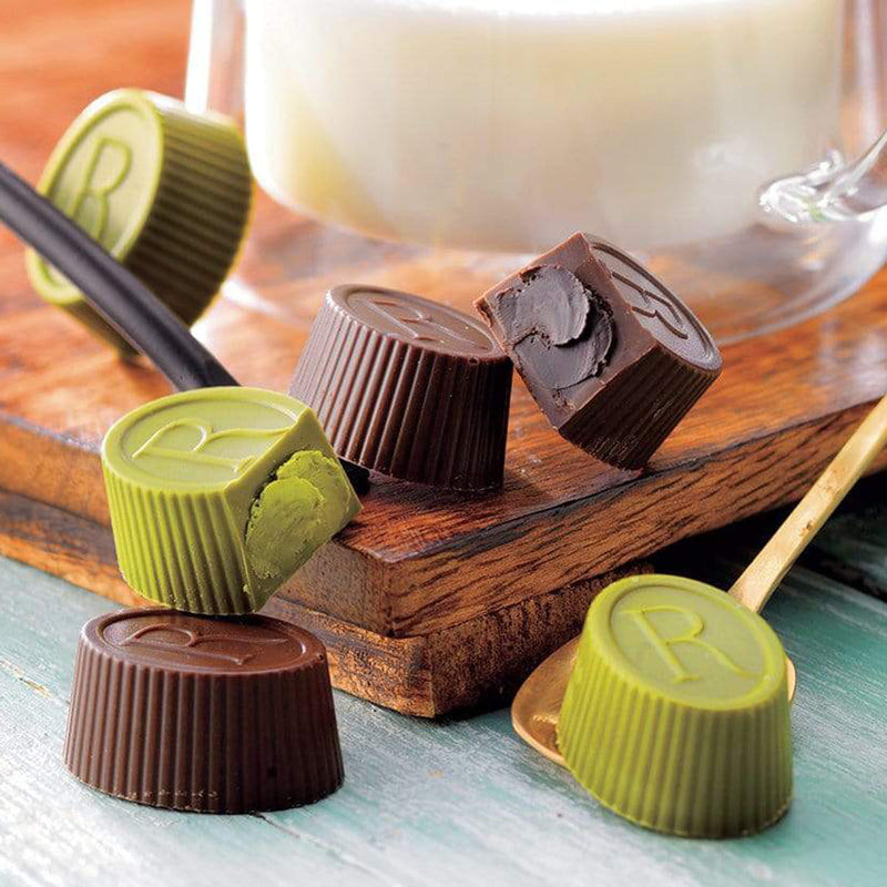 ROYCE' Chocolate - Tea Chocolat Hojicha & Matcha - Image shows brown and green chocolate cups engraved with the letter R. Accents include brown wooden trays and spoons. Background features a glass of milk and a wooden surface in color blue.