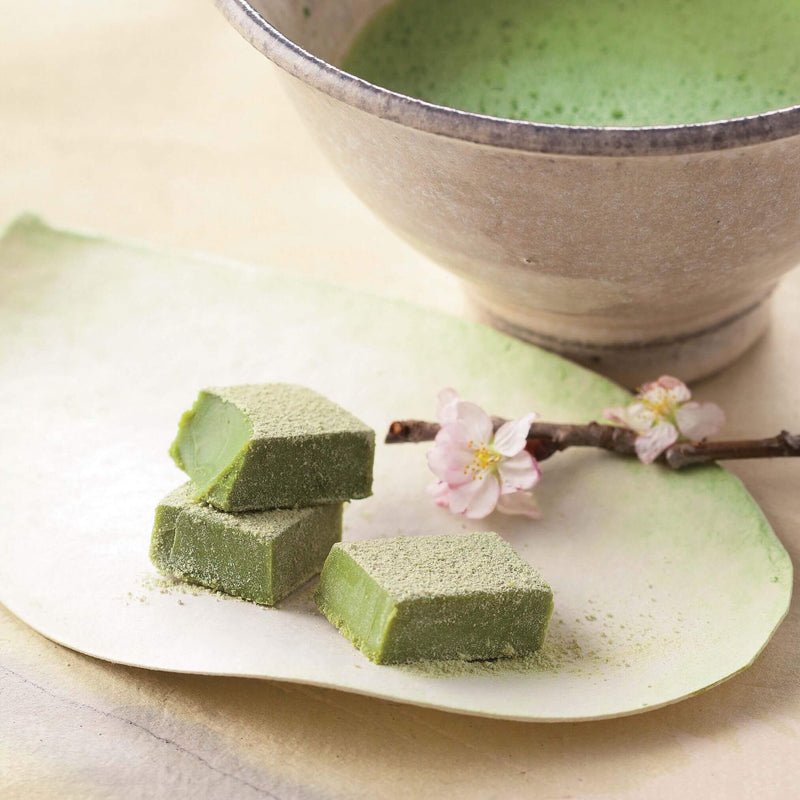 ROYCE' Chocolate - Nama Chocolate Matcha - Image shows green blocks of chocolates on a plate. Accents include a brown stem with pink flowers and a white tea cup filled with green tea.