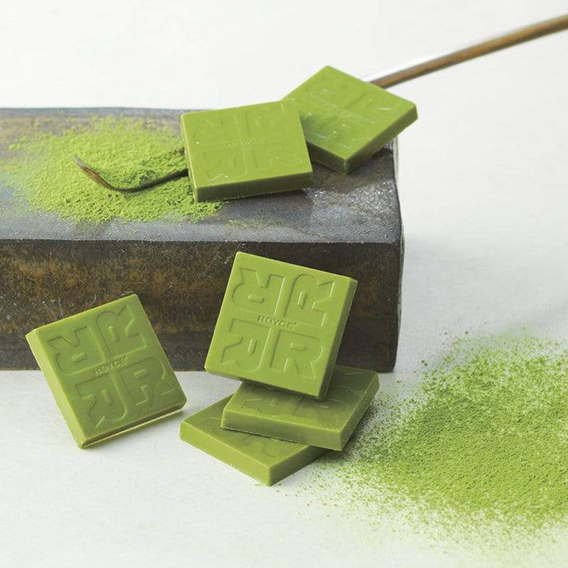 ROYCE' Chocolate - Matcha Chocolate - Image shows green chocolate squares etched with R letters and the word ROYCE. Accents include a brown wooden stick, green tea powder, and a gray stone platform. Background is white.