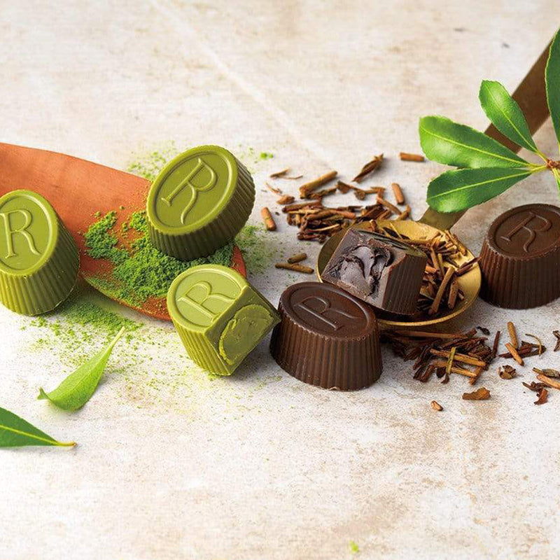 ROYCE' Chocolate - Tea Chocolat Hojicha & Matcha - Image shows brown and green chocolate cups engraved with the letter R. Accents include wooden cups, green leaves and powder, and brown tea shavings. Background and surface is light brown.