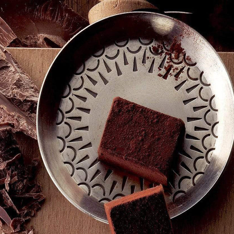 ROYCE' Chocolate - Nama Chocolate Mild Cacao - Image shows brown blocks of chocolates on a silver metallic plate. Accents include a brown wooden platform under and brown chocolate shavings on the side.