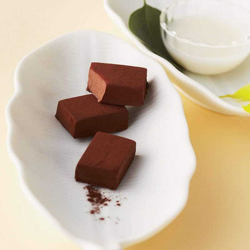 ROYCE' Chocolate - Nama Chocolate Mild Cacao - Image shows brown blocks of chocolates on a white leaf-shaped plate. Accents include another white leaf-shaped plate with a bowl of milk and green leaves. Background is in light brown.